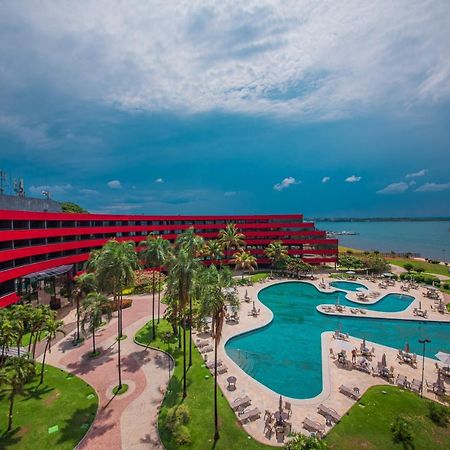 로열 튤립 브라질리아 알보라다 호텔 외부 사진 The photo shows a modern hotel building with a striking red exterior, surrounded by lush greenery. In the foreground, there is a large, creatively shaped swimming pool, with lounge chairs arranged around it. The backdrop features a body of water, pos