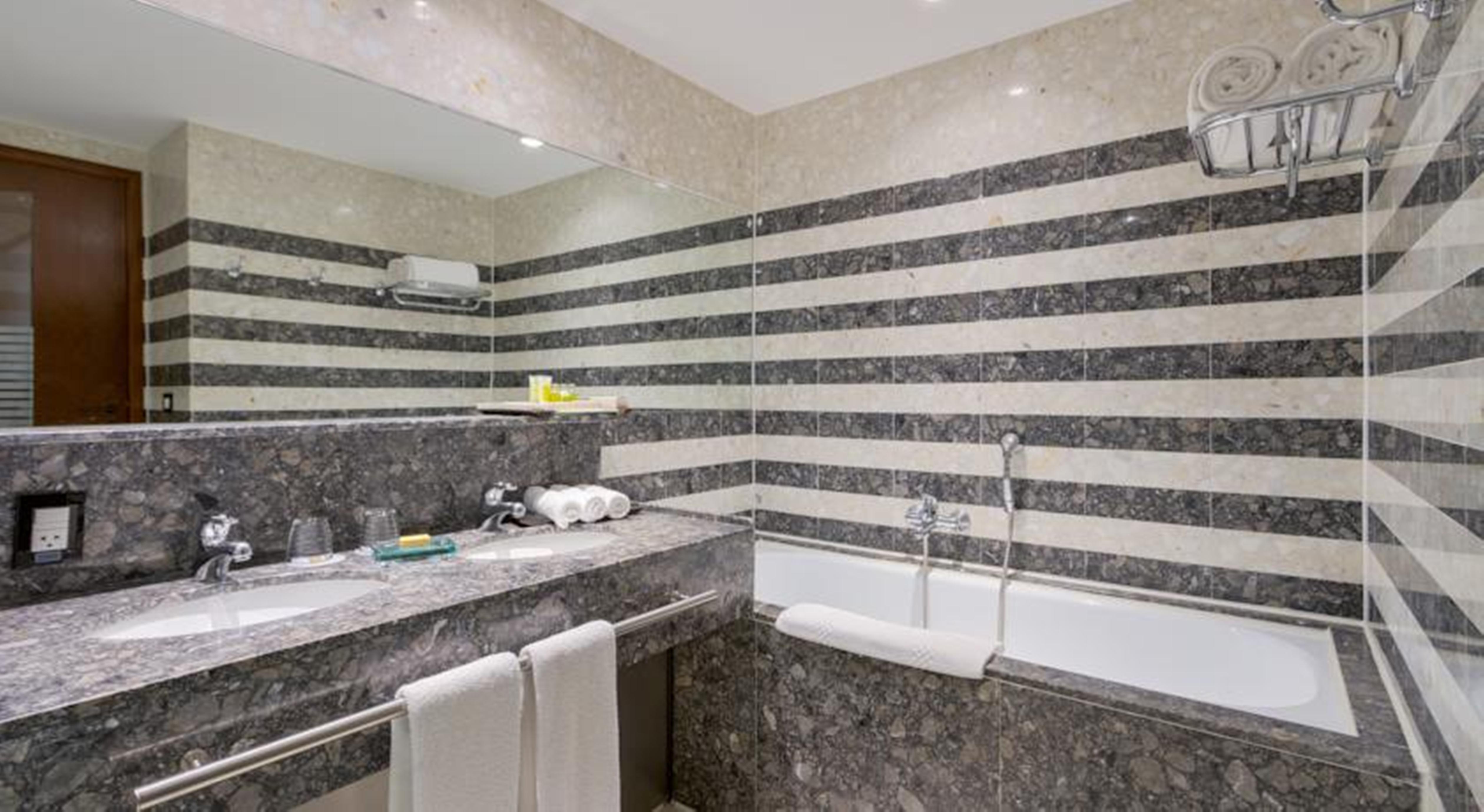 로열 튤립 브라질리아 알보라다 호텔 외부 사진 The photo shows a modern bathroom featuring a combination of marble and stone finishes. The walls are decorated with horizontal stripes in various shades of gray and cream. There is a bathtub with a white exterior, and a showerhead is mounted on the 