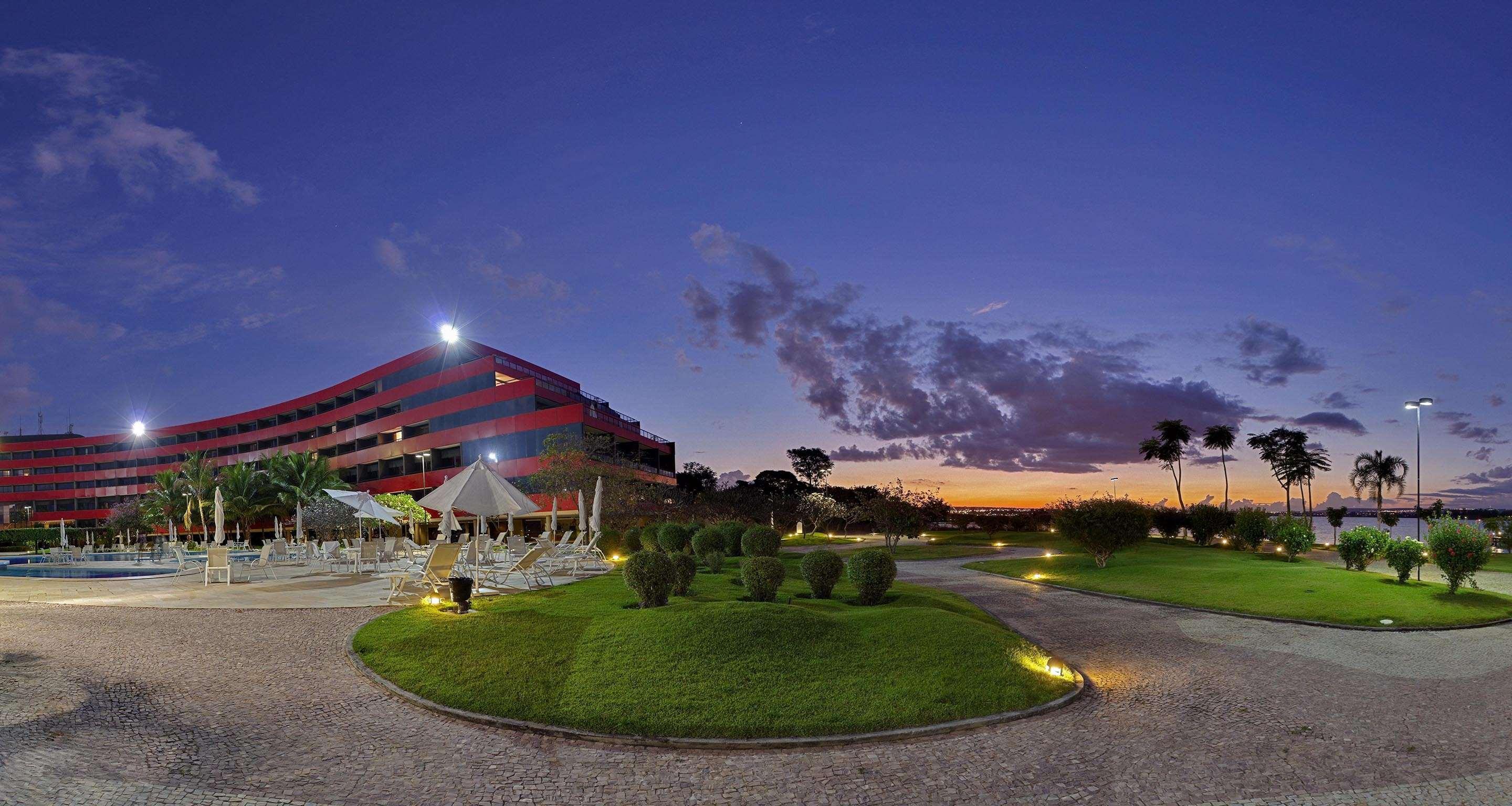 로열 튤립 브라질리아 알보라다 호텔 외부 사진 The photo depicts a modern building, possibly a hotel or resort, with a striking red exterior. It is set in a landscaped area that features well-maintained greenery and ornamental plants. In the foreground, there are lounge areas with umbrellas, sugg