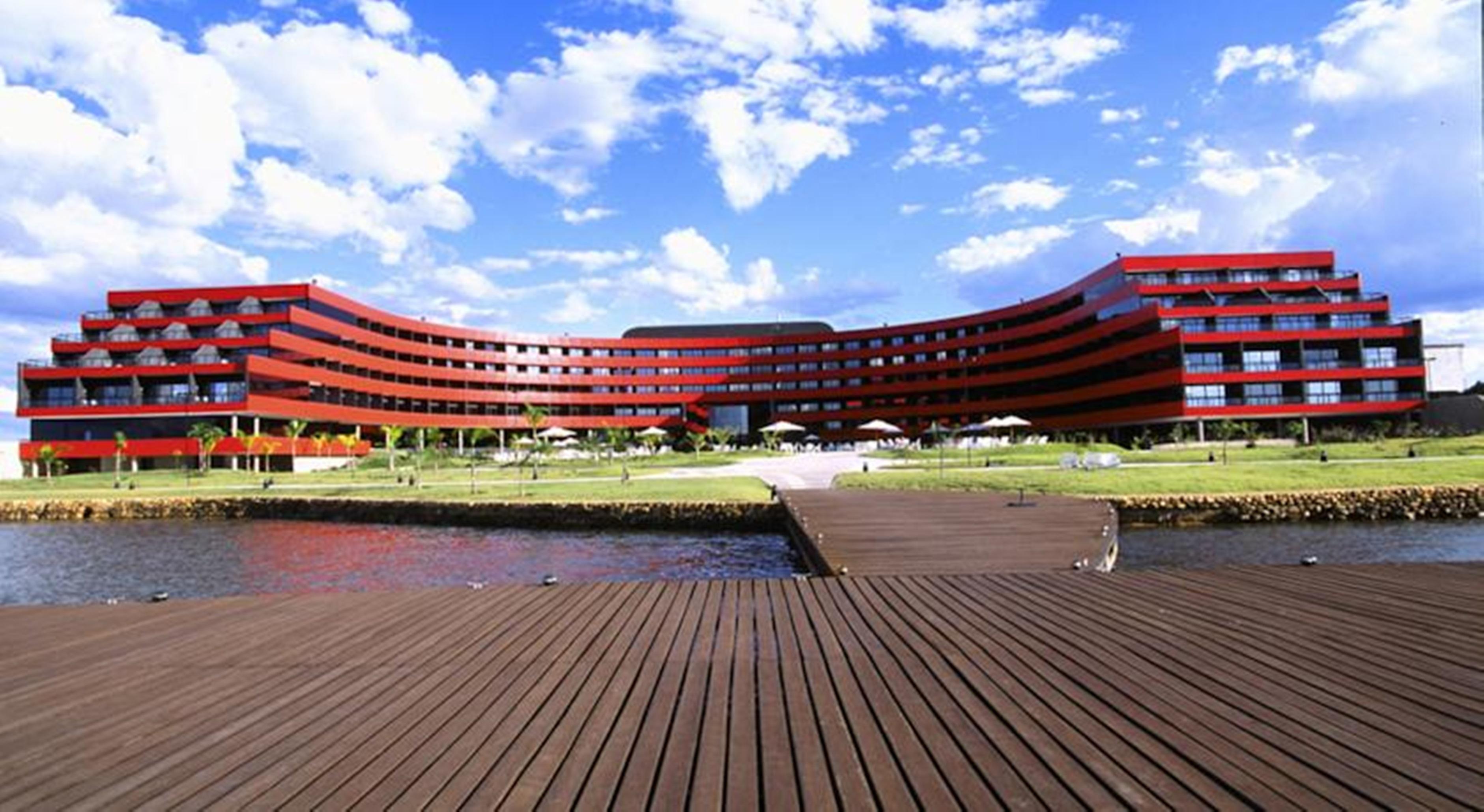 로열 튤립 브라질리아 알보라다 호텔 외부 사진 The image shows a modern, curved building with a striking red facade, situated near a body of water. The architecture features multiple levels with large windows that create a sleek and contemporary appearance. In front of the building, there is a wo