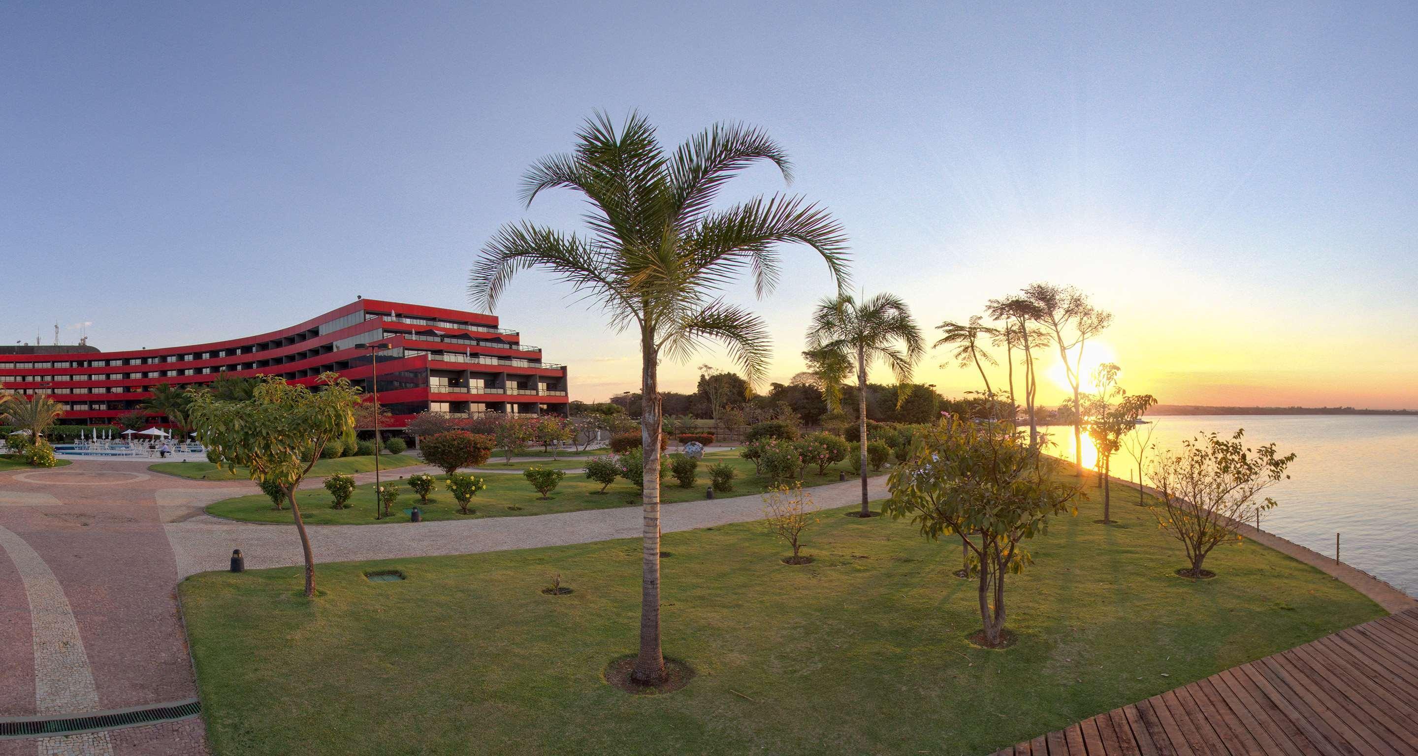 로열 튤립 브라질리아 알보라다 호텔 외부 사진 The photo depicts a scenic view featuring a red multi-story building alongside a serene waterfront. In the foreground, there are several palm trees and lush green lawns, creating a tropical atmosphere. The sun is setting in the background, casting wa