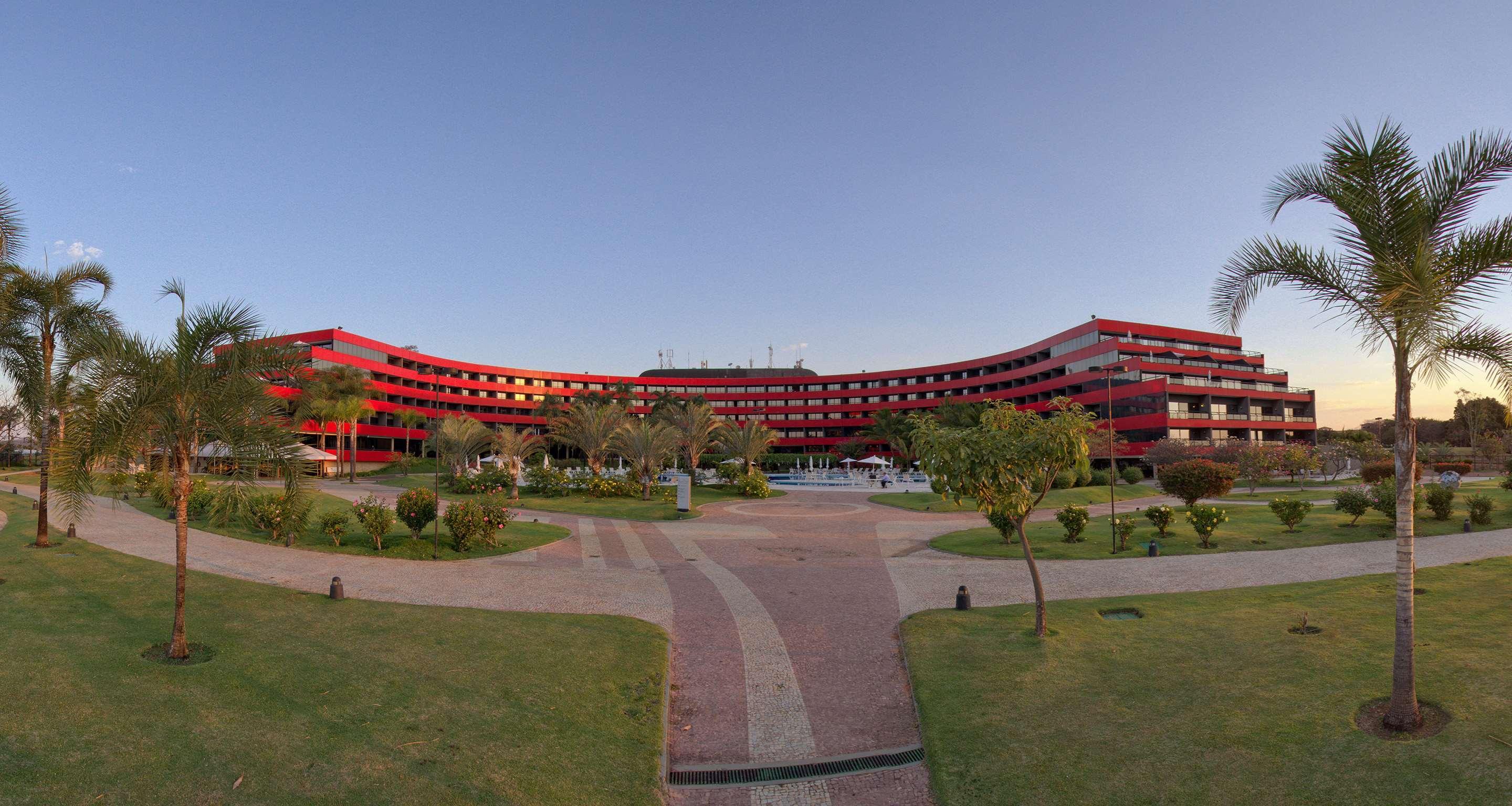 로열 튤립 브라질리아 알보라다 호텔 외부 사진 The photo shows a large, curved building with a vibrant red exterior. It appears to be a hotel or resort, surrounded by well-maintained gardens and green spaces. The landscape includes manicured lawns, small trees, and pathways, suggesting a pleasant