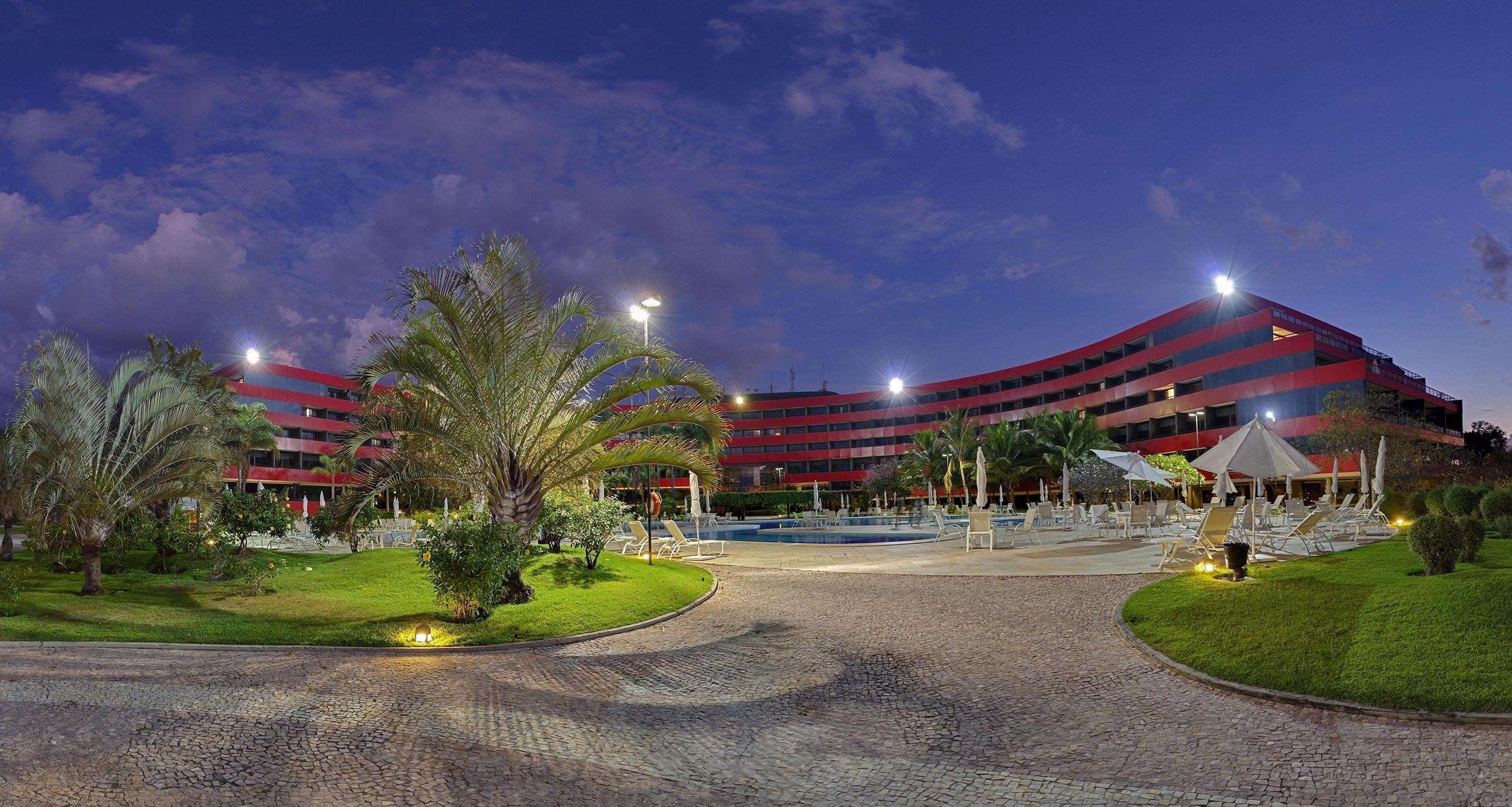 로열 튤립 브라질리아 알보라다 호텔 외부 사진 The photo shows a panoramic view of a hotel or resort complex, characterized by its modern architectural design with smooth, curved lines and a vibrant red color. It’s set in a landscaped area featuring palm trees and green grass. The surroundings in
