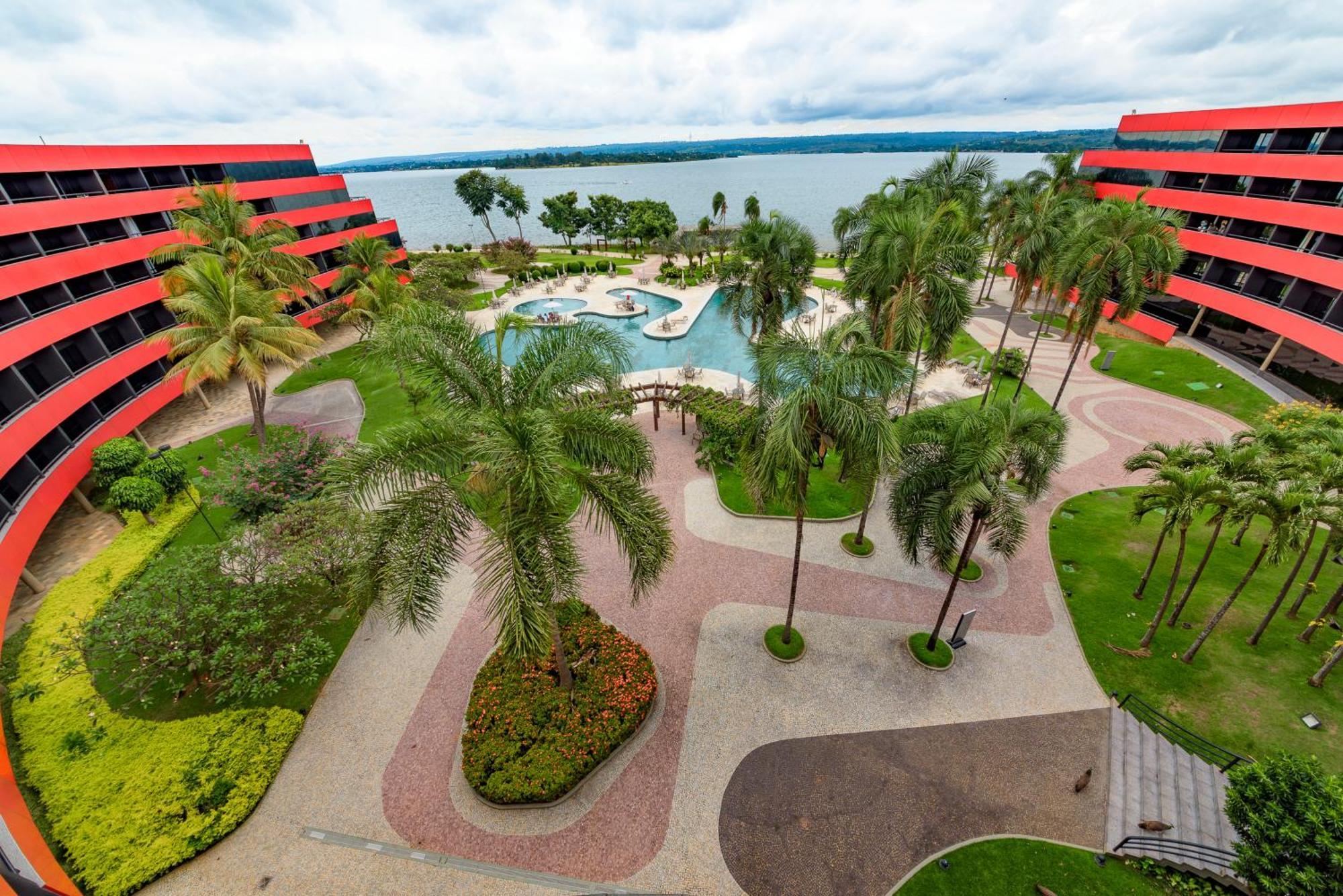 로열 튤립 브라질리아 알보라다 호텔 외부 사진 The photo shows a scenic view of a courtyard area in a residential complex. There are multiple red buildings surrounding a pool area, which is surrounded by palm trees and landscaped gardens. The pool features a curvy design, and there are pathways m