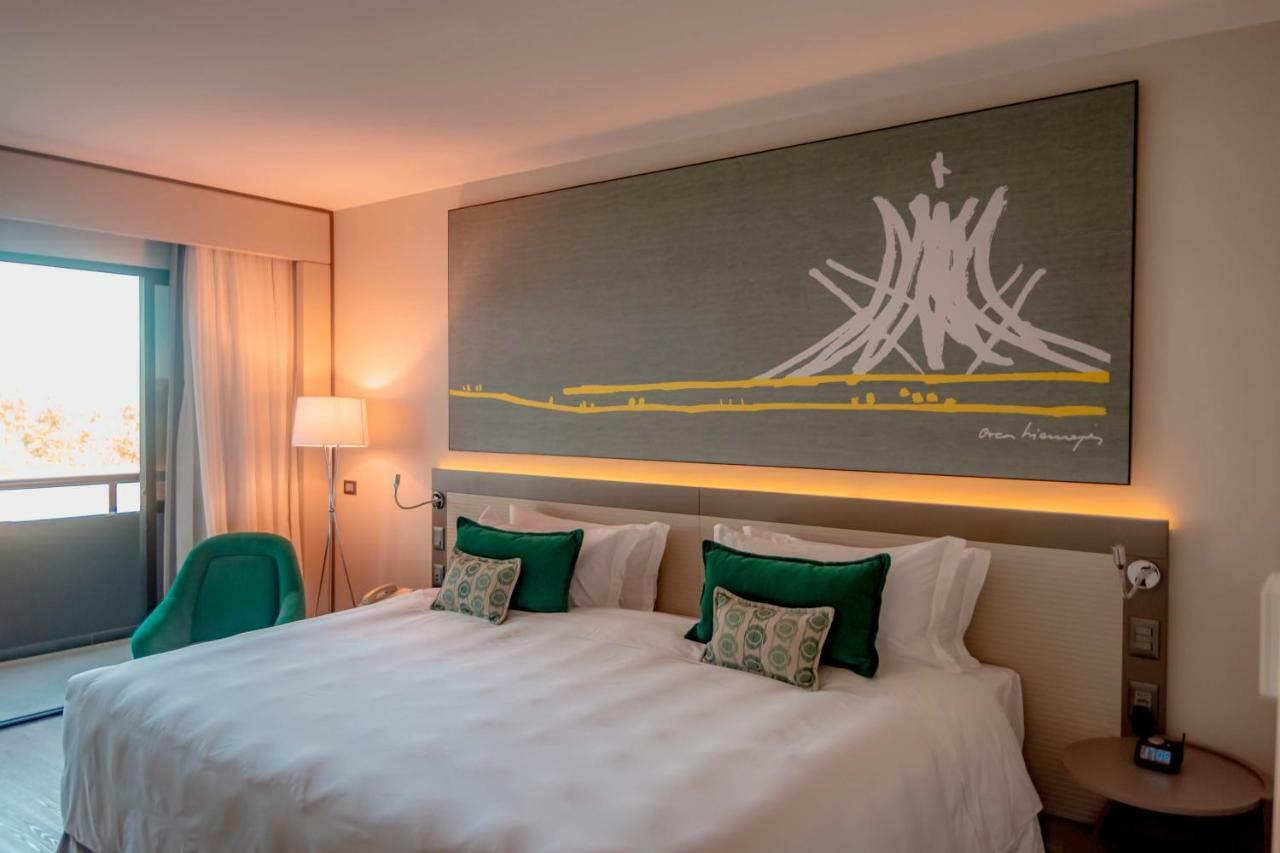 로열 튤립 브라질리아 알보라다 호텔 외부 사진 The photo shows a modern hotel room featuring a large bed with white bedding and two green accent pillows. Above the bed, there is a large piece of artwork depicting a stylized mountain or volcanic shape with yellow accents. The room has a contempora